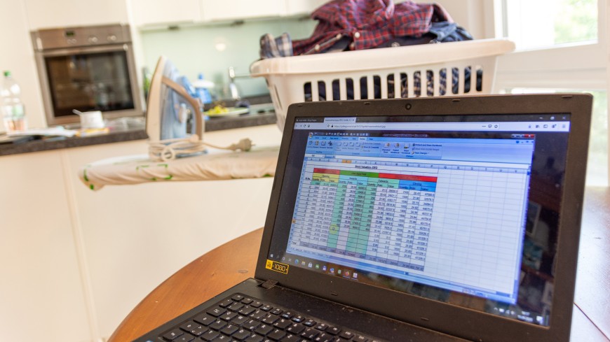 Ein Laptop mit einer Excell-Tabelle steht im Vordergrund, im Hintergrund sieht man Bügelwäsche in einer Küche.