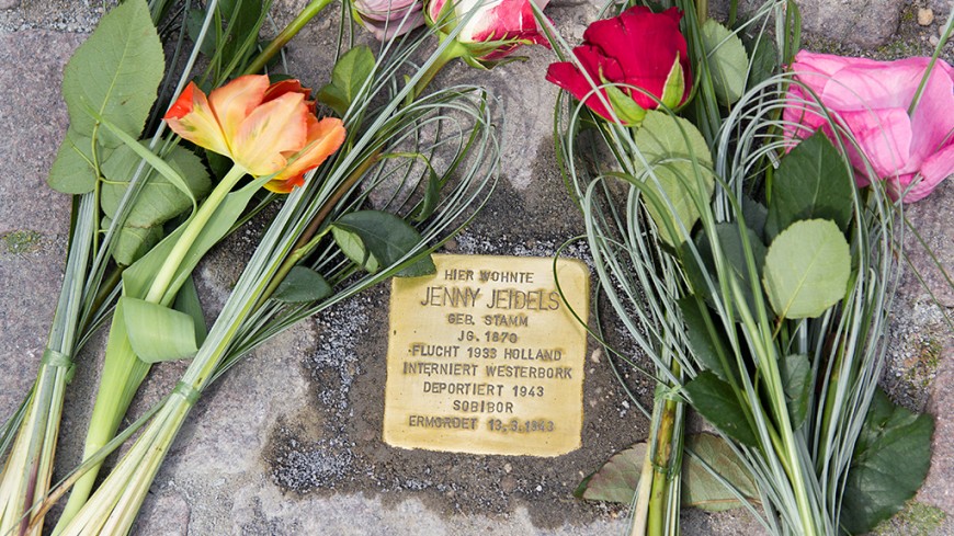 Nahaufnahme eines "Stolpersteins" mit Blumen drumherum. Bild: Paul Glogowski