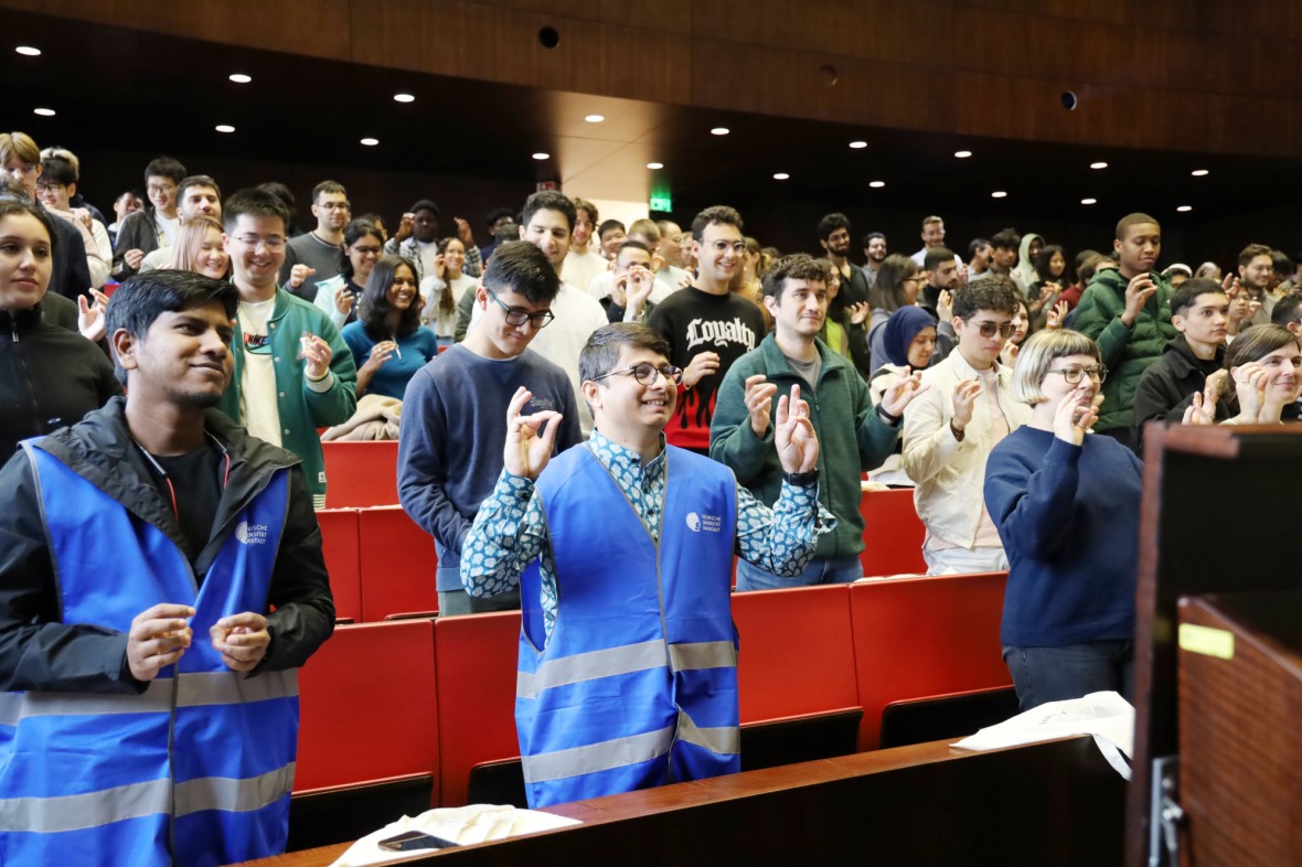 Das Foto zeigt ein lebhaftes Willkommensevent an der TU Darmstadt. Eine große Gruppe ist in einem Vorlesungsaal mit dunklen Holzwänden und roten Stühlen. Die meisten Personen stehten und halten ihre Hände in die Höhe und machen Fingerübungen. in der ersten Reihe Stehen Studierende mit blauen Warnwesten. Das sind MasterPlus-Buddys, die Masterstudierende betreuen.