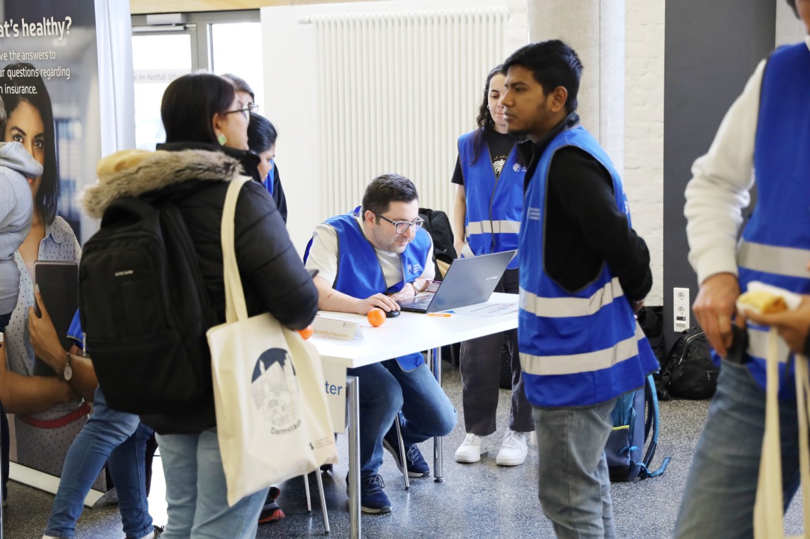 Infomesse während der Willkommensveranstaltung Kick-Off Days. Einige Studierende tragen blaue Westen, arbeiten dabei am Laptop an einem Tisch umgeben von anderen Personen in blauen Westen und beraten neue Masterstudierende. Die Personen mit blauen Westen sind MasterPlus Buddys.