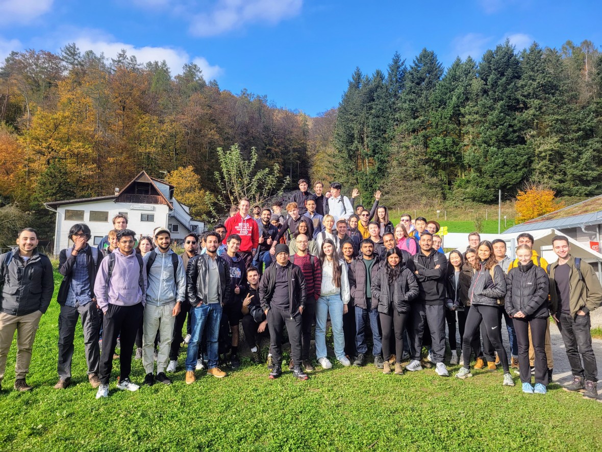Auf dem Gruppenfoto sind über 50 junge Studierende bei einem Ausflug in der Natur zu sehen. Sie stehen auf einer Grünfläche. Hinter ihnen sind viele Bäume zu sehen. Einige Laubbäume haben bereits gelbe Blätter. Die Sonne scheint und sie blicken fröhlich in die Kamera. Zu sehen ist ein ESN-Ausflug, an dem die Masterplus-Buddys mit den Masterstudierenden teilnehmen, die sie betreuen.
