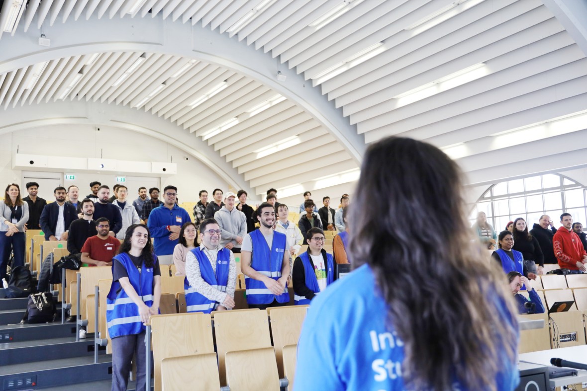Ein heller Vorlesungssaal, gefüllt mit fröhlichen Menschen. Die meisten Personen stehen und lächeln. In der ersten Reihe tragen junge Studierende blaue Westen. Sie sind MasterPlus Buddies und nehmen an der Willkommensveranstaltung der Kick-Off Days teil, um neue internationale Studierende zu begrüßen. Im Vordergrund ist eine Person von hinten unscharf zu sehen.