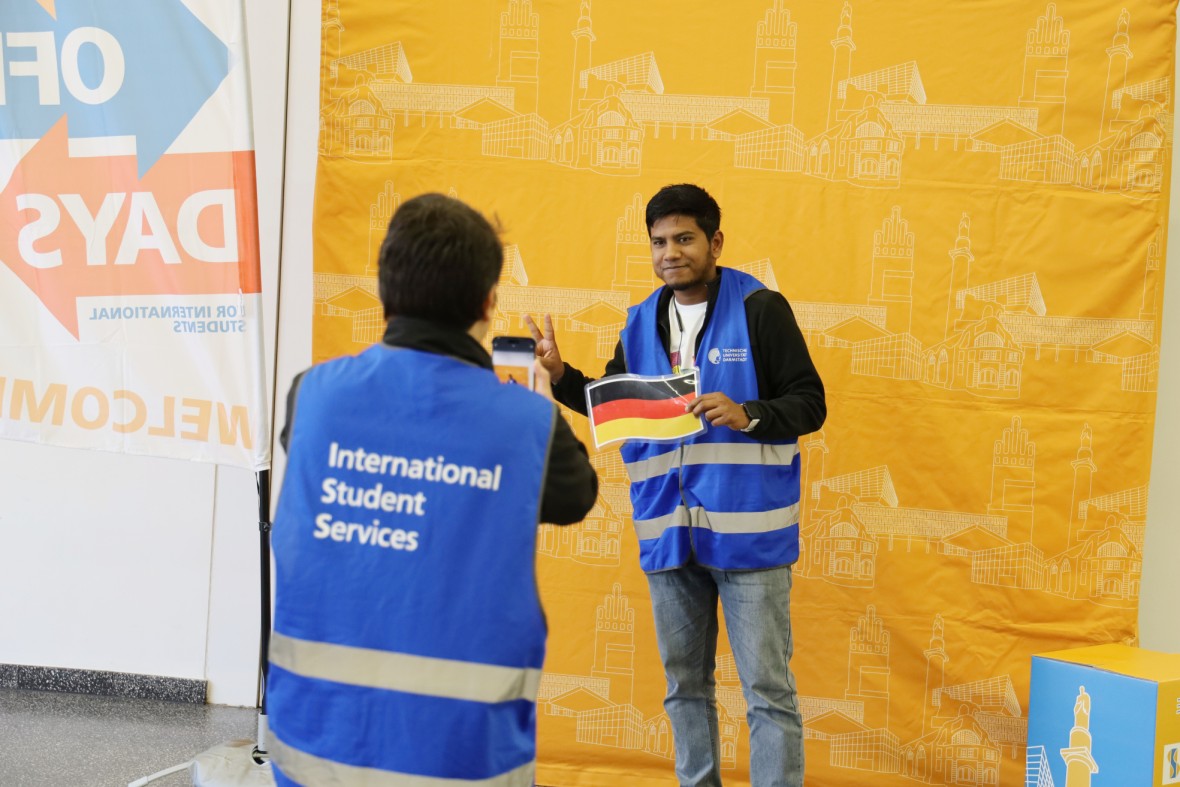 The image shows two people, both wearing blue high-visibility vests. They are MasterPlus buddies. One of them is holding a small German flag in front of them and posing smiling for a photo, while the other person is taking a picture with their smartphone. An orange background can be seen in the background. On the left is a beach flag indicating that this is the ‘Kick-Off Days’ event.