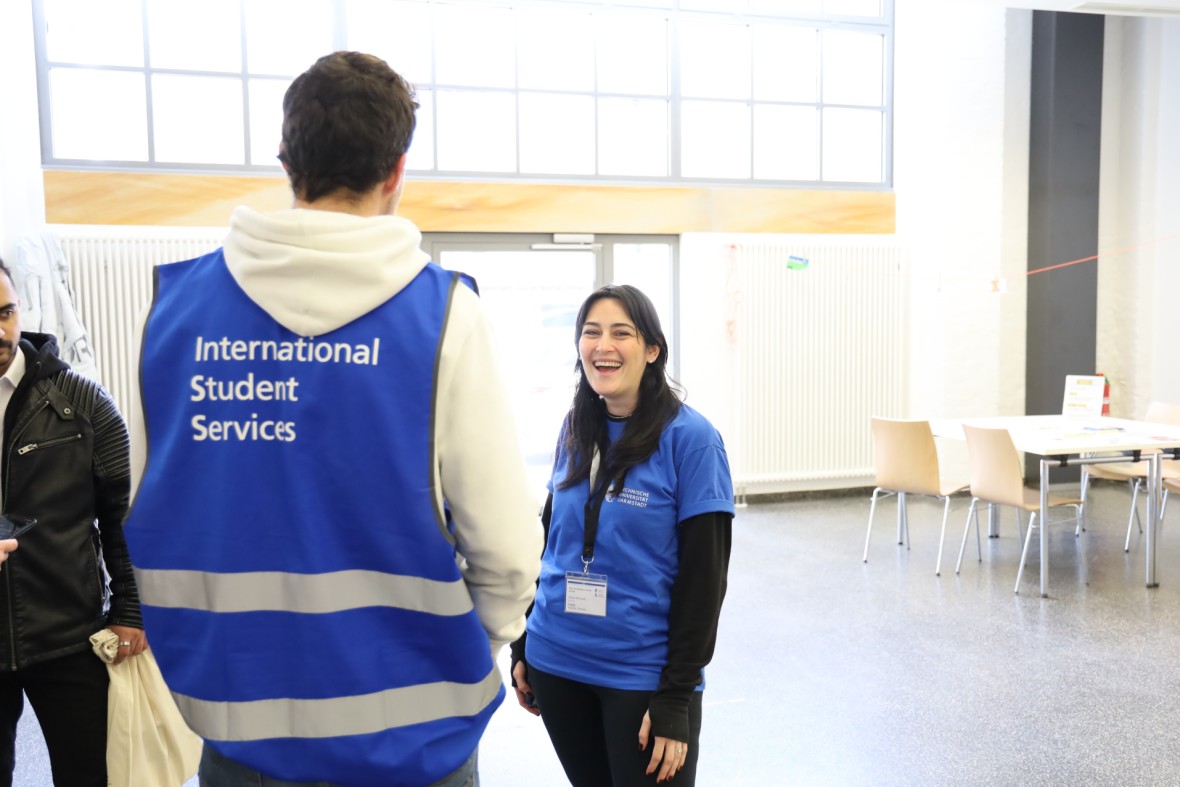 The back of a MasterPlus buddy can be seen, recognisable by his blue ISS vest. In front of him, a woman wearing a blue ISS T-shirt smiles at the camera.