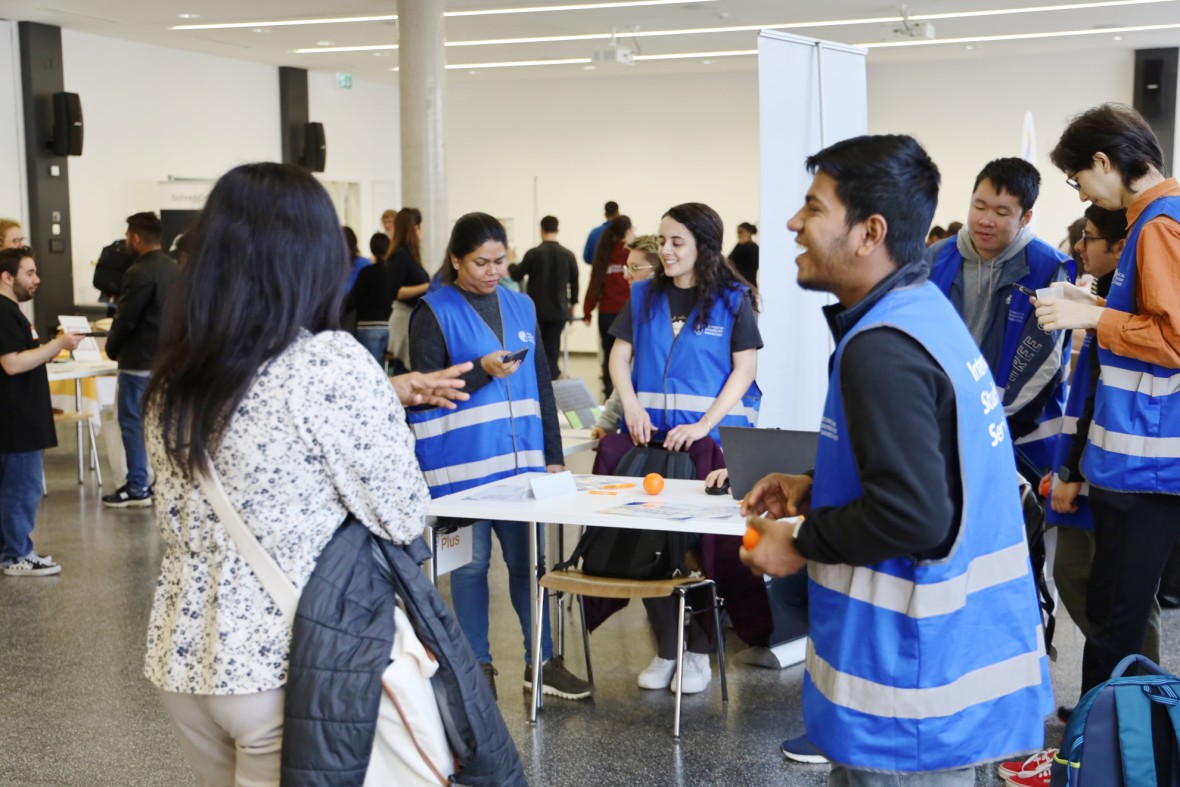 The MasterPlus Buddies are standing around a desk in their blue vests, talking and laughing. At the front, a Buddy is laughing with a student.