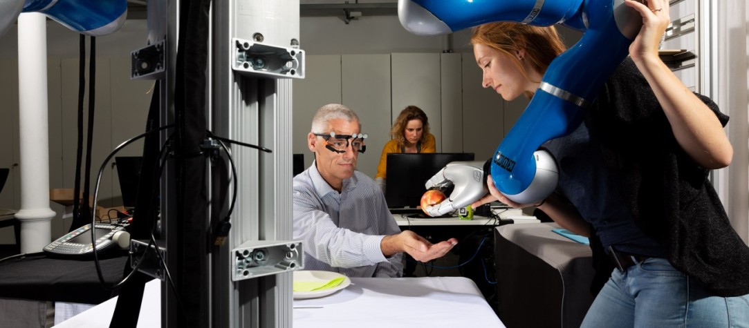 Eine Studentin der TU Darmstadt bedient eine großen Roboterarm. Dieser reicht einem Mann einen Apfel. 