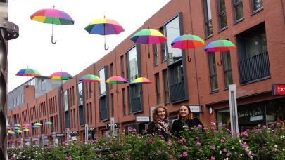 Zwei Frauen stehen auf einer blumengeschmückten Brücke, unter Regenbogenschirmen / Two females standing on the bridge decorated with flowers, under rainbow umbrellas