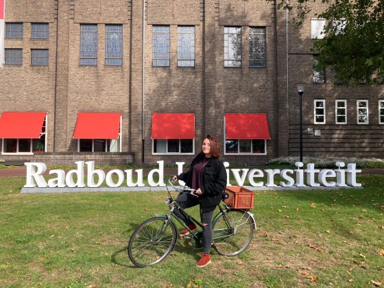 Eine Studentin auf einem Fahrrad steht vor dem Schriftzug der Radboud University in Nijmegen vor einem Universitätsgebäude.