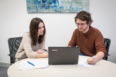 Beratungssituation in einem Mentoring-Kontext zweier Studierender an einem Schreibtisch mit Laptop und Schreibmaterialien. // Counselling situation in a mentoring context of two students at a desk with laptop and writing materials.